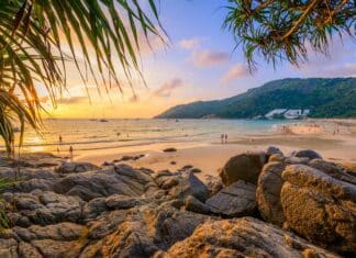 Coucher de soleil à Nai Harn Beach, Phuket, avec un ciel orange, des nageurs, des personnes sur la plage sablonneuse, des voiliers en mer et des rochers le long du littoral.